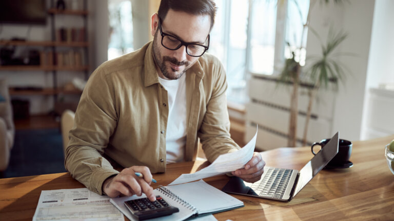 Homme sur son ordinateur qui calcule et tient des papiers en main